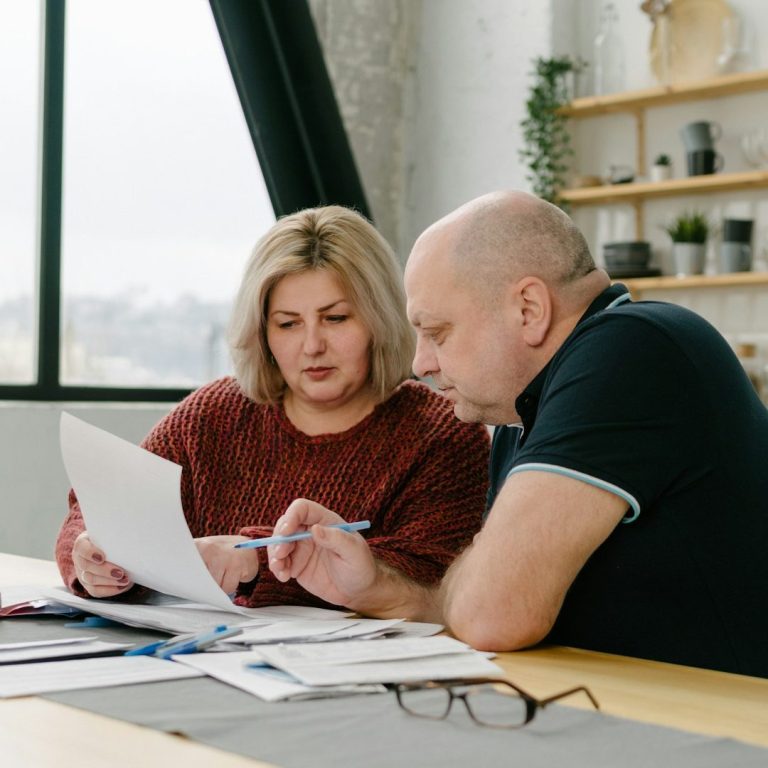Zwei Personen am Tisch, die Dokumente prüfen und besprechen. Grünpflanzen im Hintergrund.