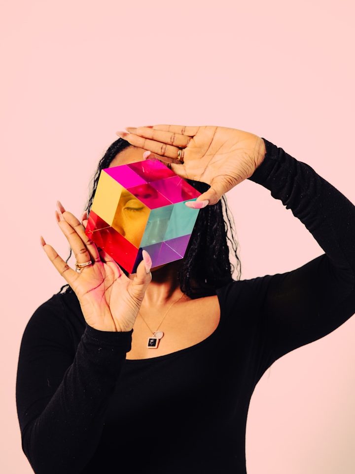 Eine Person hält ein buntes, geometrisches Objekt vor ihr Gesicht vor einem rosa Hintergrund.
