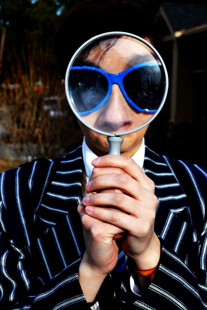 Person in gestreiftem Anzug und blauen Sonnenbrillen hält eine Lupe vor dem Gesicht.